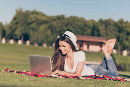 Young happy girl is typing in her laptop lying on the plaid on grass at park in spring outdoorsの写真素材