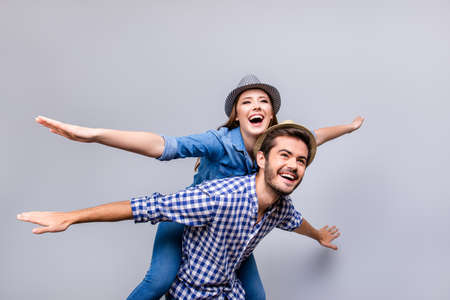 Cheerful and playful couple in casual outfits and hats are fooling around, gesturing plane wings, smiling, posing, going crazy on pure light backgroundの写真素材