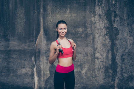 Happy young sport woman in stylish training outfit with jumping rope on her shoulders is smiling, standing outside on a concrete wall`s backgroundの写真素材