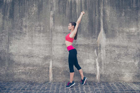 Young sportwoman trainer is making stretching outdoors in town. She is so relaxed and healthy, wearing modern pink and black sport outfit, on the concrete wall`s backgroundの写真素材