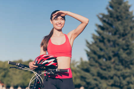 Cute young sporty lady is outside on a summer day, finished cycling on modern bike, in fashionable outfit, with helmet, looking farの写真素材