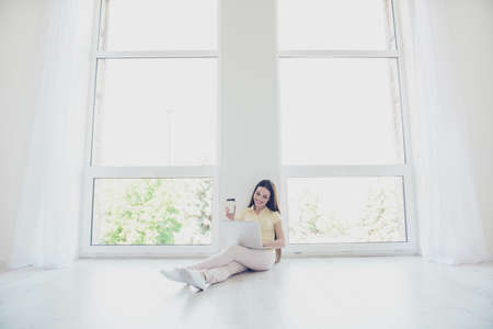 Young happy girl is typing on her laptop sitting on the floor indoors at home, having tea, smilingの写真素材