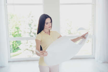 Focused young pretty designer is looking at the scheme of the project, she is in a casual outfit, with long hair, standing in light roomの写真素材