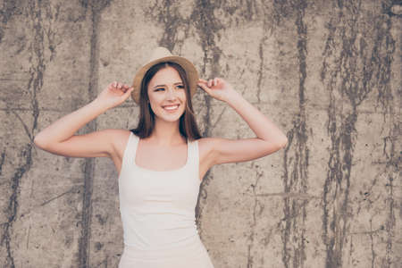 Happy young girl on summer vacation. She is in a stylish hat, wearing casual light dress, holding her beige cap, amazed, on concrete wall`s background, walking outdoorsの写真素材