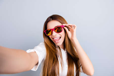 Attractive young lady is making a selfie on the camera, flirty and playful. In pink trendy sunglasses, with tongue out, on pure light grey backgroundの写真素材