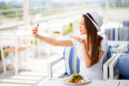 Profile side photo of young girl, having salad on a summer open air light terrace outside. She is in a casual wear, hat, taking selfie, sending an air kissの写真素材