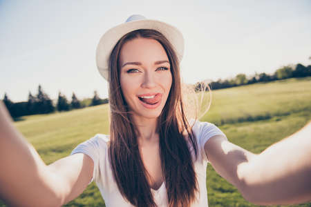 Happy young girl on vacation in an open air spring park is making selfie, wearing casual outfit, hat, posing with tongue outの写真素材