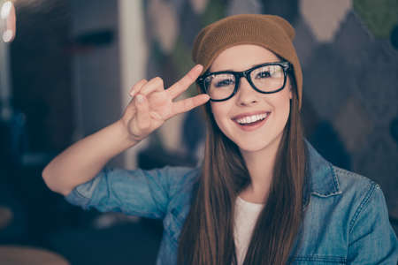 Close up portrait of cheerful playful girl, who is gesturing peace sign and smiles. She is wearing casual clothes, glasses and a brown hatの写真素材