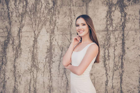 Young slim cute girl in white elegant dress, is so successful and beautiful, tempting pose. Behind her is a concrete wall, she is walking outdoors in the cityの写真素材