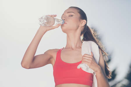Close up low angle photo of young fit sportwoman, she finished her work out and now drinking water to refresh. She is outside on a summer stadium, with a towelの写真素材