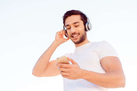 Smiling muscular young brunet is enjoying listening to the music in modern headphones, isolated on white background, in white tshirt, on his mobile phoneの写真素材