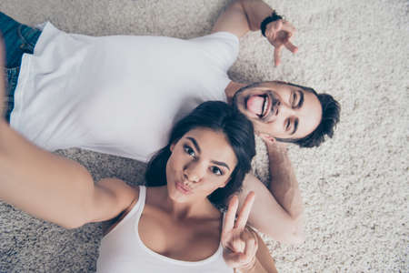 Cheerful playful ltino mulatto sister and brother are taking selfie and making funny grimaces, gesturing. They lie on the floor on beige carpet in white casual outfits indoors at homeの写真素材