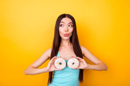 Sexy young attractive korean is holding sweet donutes near her breasts, standing on bright yellow background, in blue casual outfitの写真素材