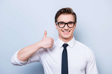 Successful excited young guy in glasses and formal wear is standing isolated on a pure background, showing thumb up sign, smilingの写真素材