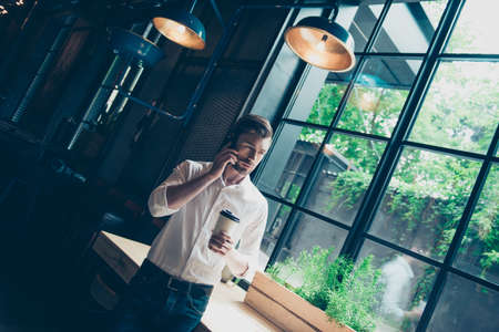 Successful young man, stands near window with view on the garden, has a business conversation,  at work station in a modern coworking, well dressed, looking awayの写真素材