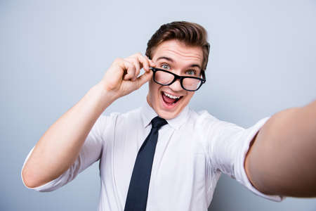 Funky mood of an excited geek young man in glasses and formal wear. He is making selfie shot on camera, standing on a pure background, fooling aroundの写真素材