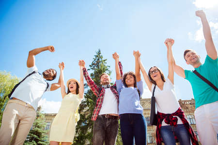 Low angle view of happy students with raised arms together, wearing casual clothes, walking after studies outside in a sunny spring day. Conception of successful teambuildingの写真素材