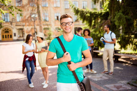 Young successful blond nerdy student is standing with bag and smiles, behind are his classmates, park near campus, sunny day, carefree and enjoyable moodの写真素材