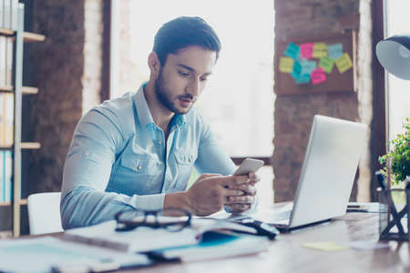 Close up of confident handsome young middle eastern lawyer guy is browsing information on his samrt phone at work placeの写真素材