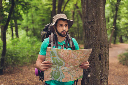 Male brunet bearded confused tourist got lost in the forest, holding map, looking far, trying to...