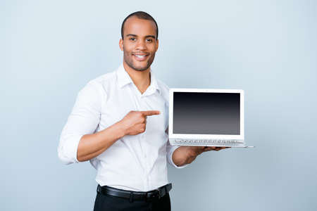Young handsome mulatto banker on the pure light blue background is pointing on a laptop`s monitor with his finger, wearing white strict shirtの写真素材