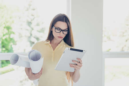 Close up of focused young pretty designer is looking at her pda, holding schemes of the project, she is in a casual outfit, with long hair, glasses, standing in light roomの写真素材