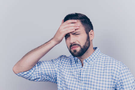 Tired sick young brunet bearded man with sad grimace and holding the forehead, wearing the checkered...