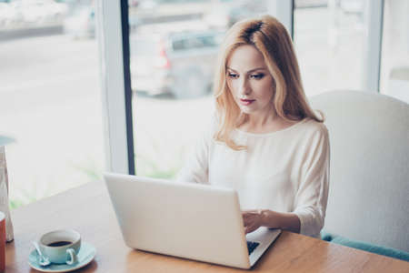Portrait of serious young blond lady entrepreneur lawyer, in white formal wear, sitting at her work place in a coworking and typing on laptopの写真素材