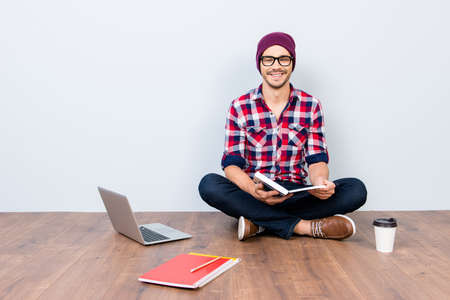 Dreamy student hipster is sitting on the floor at home with crossed legs, studying with books,...