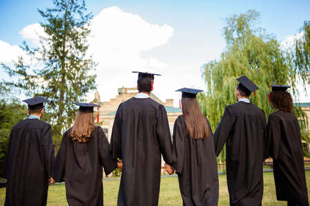 Rear view of six successful international young graduates in black robes and hats finished their education, holding hands and bonding, looking at the university building, nice sunny summer  dayの写真素材