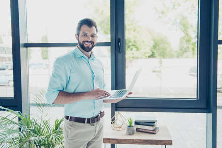 Cheerful brunet bearded business man in formal wear is typing on his laptop, he is a successful lawyer, standing at his modern light work station, smilingの写真素材