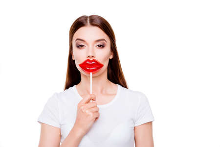 Young serious brown-haired girl is wishing to have lips like a lolipop she is holding, she stands in white outfit near the copy space, on white backgroundの写真素材