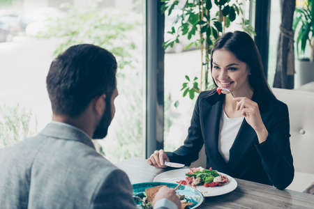 Cute young brunette lady is eating salad, looking at her partner, smiling, enjoying, in a formal suit, sitting on a terrace of a restaurantの写真素材