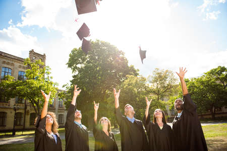 Six successful international young graduates in black robes and hats finished their education, throw mortar boards in the sky, university building behind, nice sunny summer  dayの写真素材