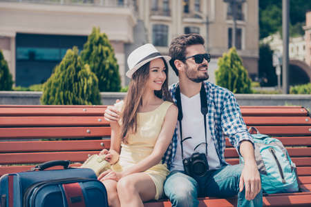 Cute married couple on vacation near the sea, sitting on bench, in casual wear, hat, spectacles, with camera and valises, smiling, loking at the viewの写真素材