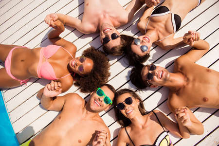Happy six international tourists on the beach in trendy sun protective wear, lying down on their backs on white wooden floor near the pool, holding hands, cheerfulの写真素材