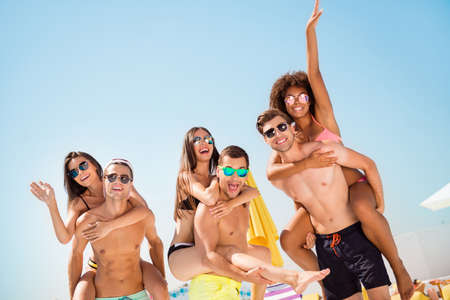 Three happy boyfriends piggybacking their girlfriends at the resort on the clear sky background, so cheerful, good day, holiday togetherの写真素材