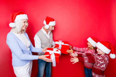 Siblings taking surprises boxes with ribbon from married elder couple of grandad and granny, in knitted cute traditional x mas clothing, enjoy, isolated on the red backgroundの写真素材