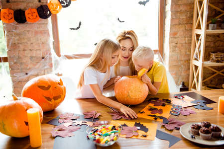 Mommy and two siblings made a jackolantern, peeking inside it, desktop with yellow candles, treats, fall leaves, little black bats on windows, garlandsの写真素材