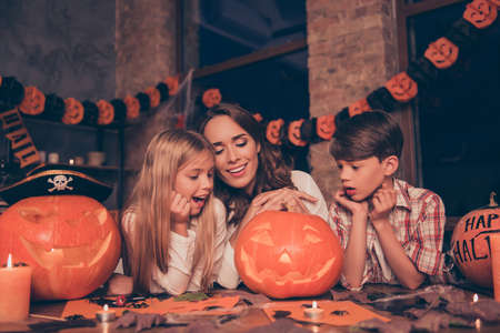 Three adorable relatives, mom with two cute small kids in decorated room, garlands on windows, looking at the cutted pumpkin, astonished and cheerful, autumn scary october feast, spooky and mysticの写真素材