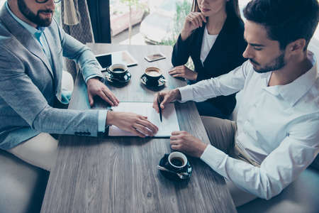 Young business partners are making a deal with lawyer about their business in a restaurant with cups of coffee. Men is signing  contract to complete the deal, reading it carefully, all are well dressed, focusedの写真素材