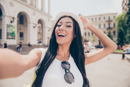 Charming mixed-race lady with bronze skin and long hair, in cap is making shot on a camera while taking a stroll outside in town. White outfit, beaming smile, posing and laughing, holds headwearの写真素材