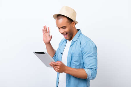 oung cheerful afro american guy is waving in camera while having video call from holiday, isolated on white background, in hat, with wide open mouthの写真素材