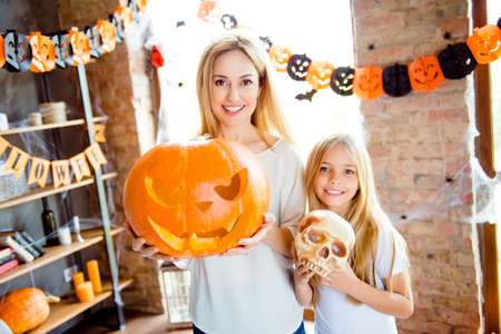 Adorable small blonde girl and her mommy are showing decorations for a halloween party, pretty mum is with big carved pumpkin, in room with garlands, bats and spider nets on the windowの写真素材