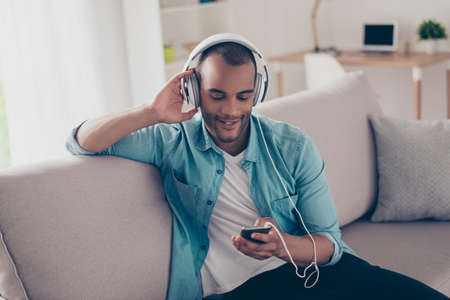 Close up portrait of serene young mulatto guy enjoying listening to nice song at home, sitting on sofa, on his smart phone, wearing casual jeans outfit, smilingの写真素材