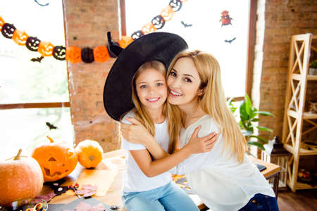 Adorable small blond girl in a black witch hat is hugging her pretty mom with such love, daughter sits on the desk top, carved pumpkins near, garlands, bats and spiders on the windows, loft styleの写真素材
