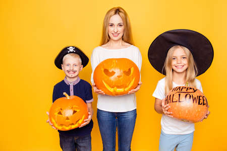 Blonde mother with two small cheerful kids, girl is a fairy, boy is a pirate, with decorations in hands, standing isolated on yellow background, with beaming smilesの写真素材