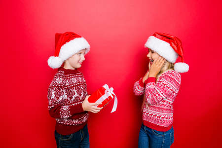Small adorable gentleman prepared present for charming smal lady, she is very excited, both in traditional knitted x mas clothing, jeans, stand isolated on red backgroundの写真素材