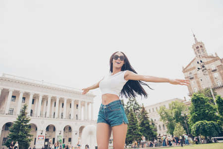 Carefree cute student in summer hot outfit is so happy, jumping outside while on stroll in town, gesturing like plane wings with hands, architecture and green trees backgroundの写真素材
