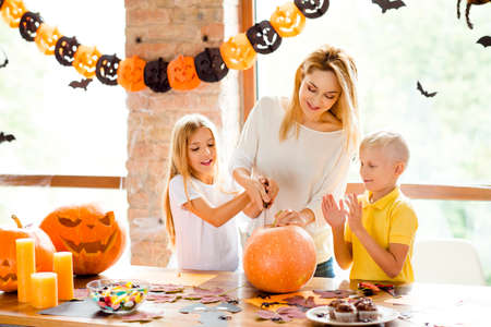 Cheerful mom with two small adorable kids preparing the jackolantern at decorated room at home loft style, desk top with yellow candles, sweets, fall leaves, little bats on windowsの写真素材